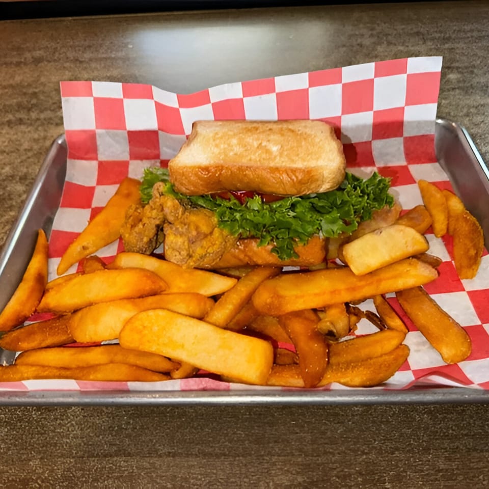 A fried chicken sandwich with lettuce and ketchup, served with golden potato wedges on checkered paper.