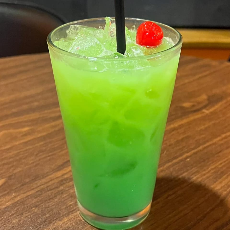 A vibrant green cocktail with ice, a black straw, and a red cherry on a wooden table.