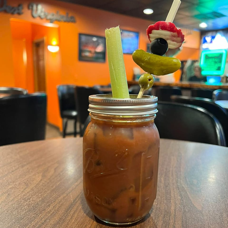 A Bloody Mary in a mason jar, garnished with celery, olives, pickle, and bacon, on a wooden table.