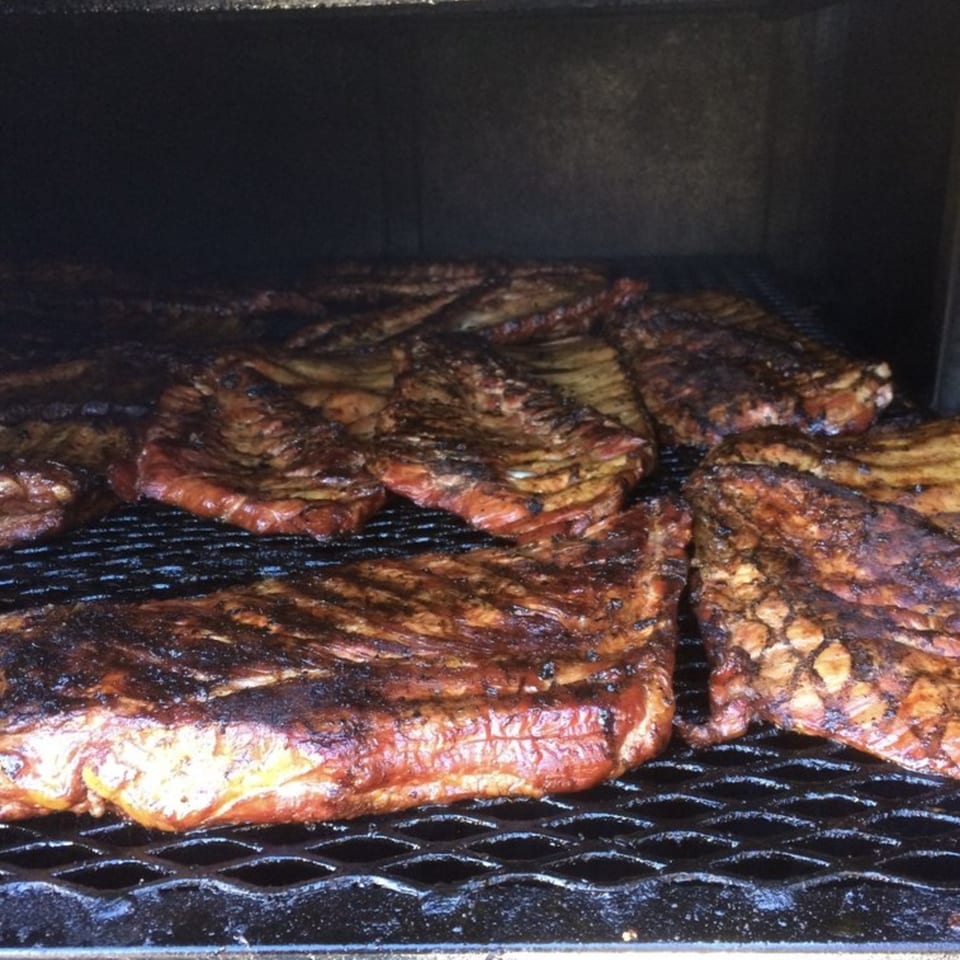 Multiple racks of Rebel SmokeHouse BBQ ribs cooking on a dark grill inside a smoker.
